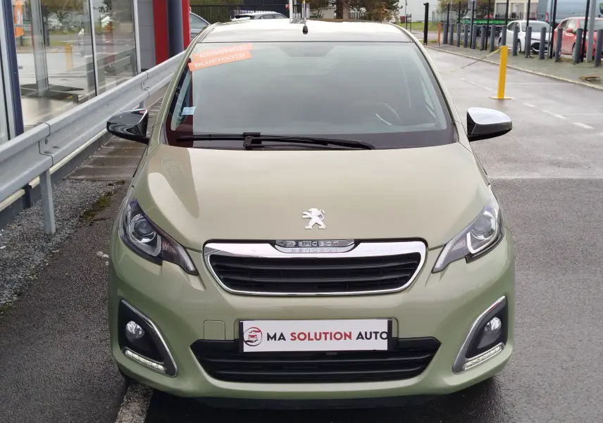 Peugeot 108 Smooth Green vue de face, avec calandre chromée et feux LED allumés, stationnée devant un bâtiment.