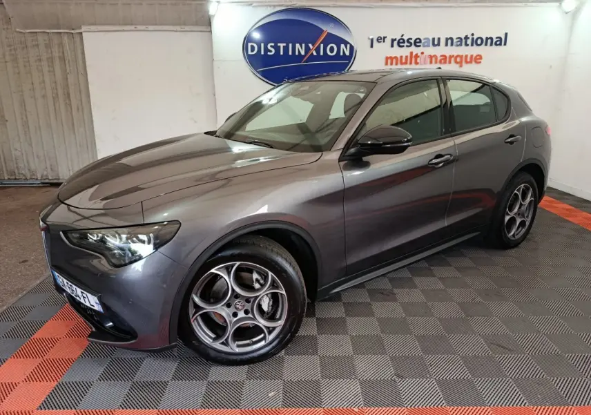 Alfa Romeo Stelvio gris foncé vu de 3/4 avant droit, avec jantes distinctives et phares LED allumés.