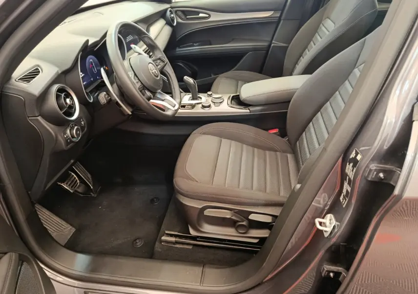 Vue côté conducteur intérieur de l'Alfa Romeo Stelvio gris foncé 2024, avec sièges tissu et tableau de bord moderne.