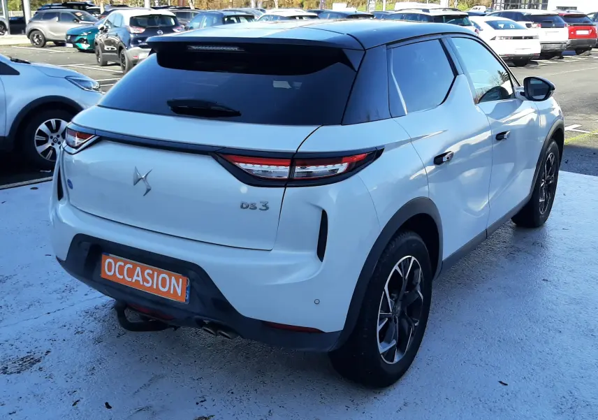 Vue 3/4 arrière droite d'un DS3 Crossback blanc perle avec toit noir et jantes noires en stationnement.