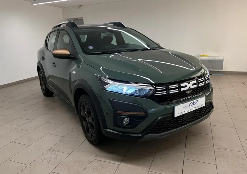 Vue 3/4 avant d'une Dacia Sandero Stepway 2023 vert cèdre avec rétroviseurs bronze et attelage visible.