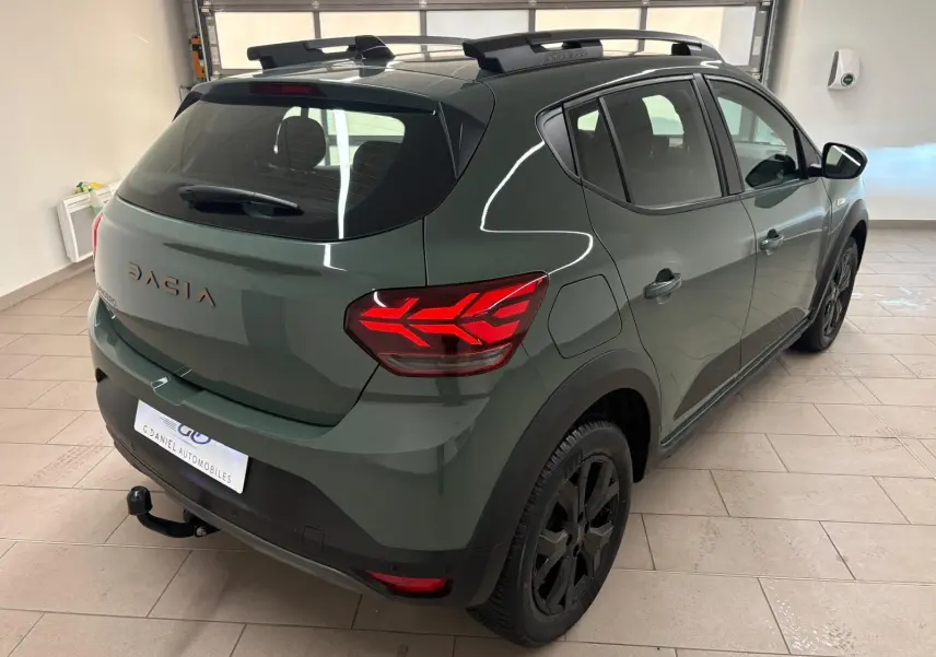 Vue 3/4 arrière droite du Dacia Sandero Stepway 2023 vert cèdre avec attelage et jantes noires en intérieur showroom.