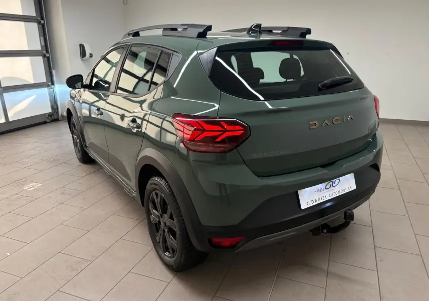 Vue 3/4 arrière droite d'une Dacia Sandero Stepway 2023 vert cèdre avec attelage et jantes noires en intérieur showroom.