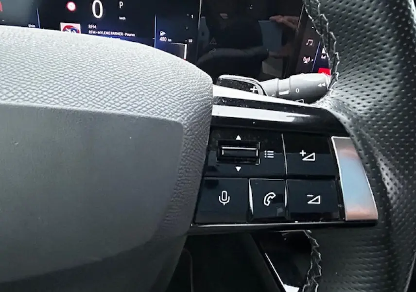 Gros plan sur les commandes au volant cuir premium de l'Opel Astra VI diesel 2022, avec affichage numérique en arrière-plan.