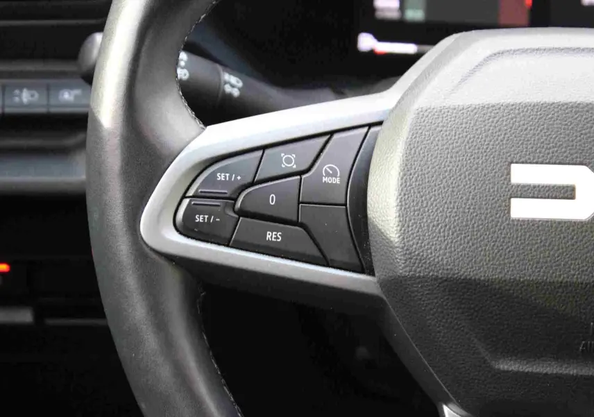 Gros plan sur le volant noir du Dacia Duster III avec commandes multifonctions côté gauche et logo partiel visible.