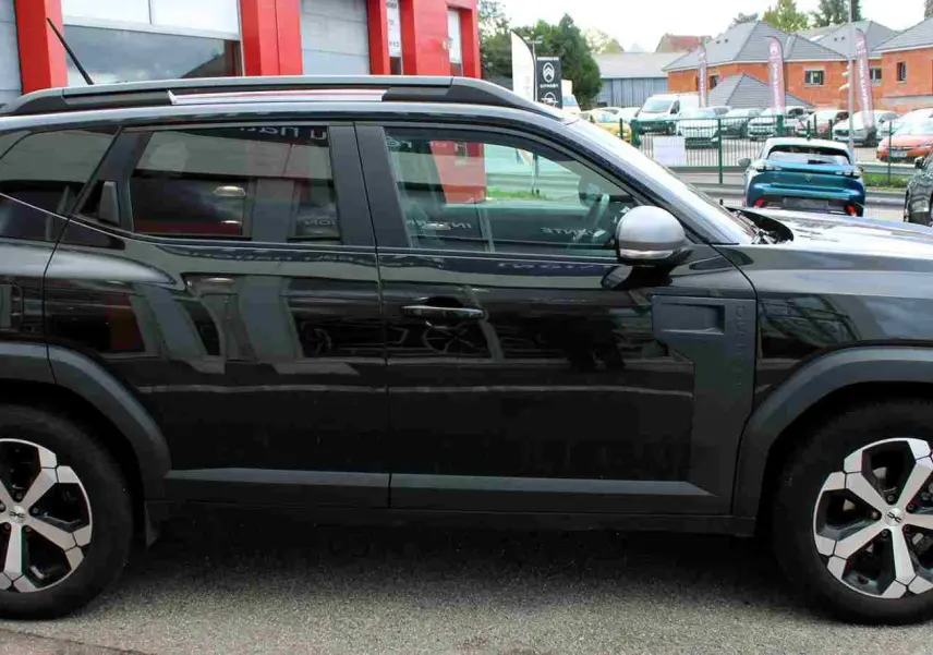 Profil droit d'un Dacia Duster noir nacré 2025 avec jantes alliage 18 pouces et barres de toit noires.