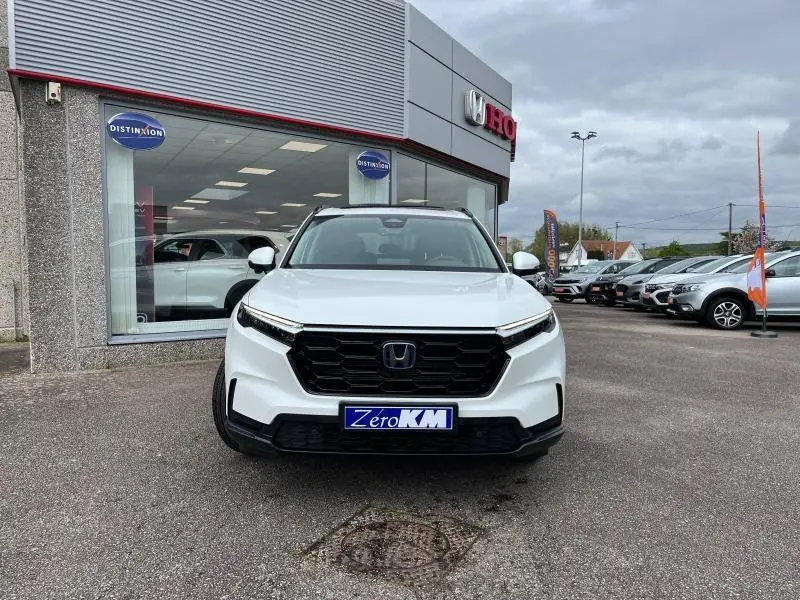 Vue de face du Honda CR-V 2024 blanc platine nacré avec calandre noire et plaque Zero KM devant un concessionnaire.
