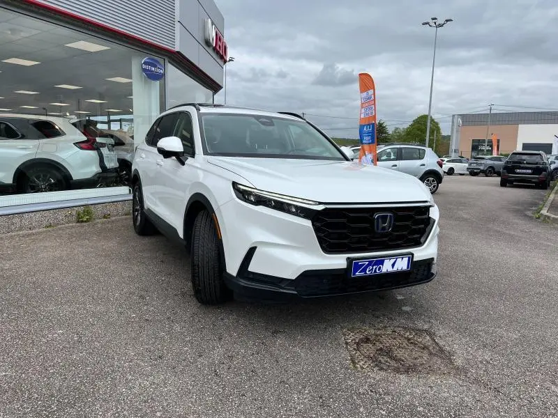 Honda CR-V blanc platine nacrée vu en 3/4 avant droit sur parking devant concession avec calandre noire distinctive.