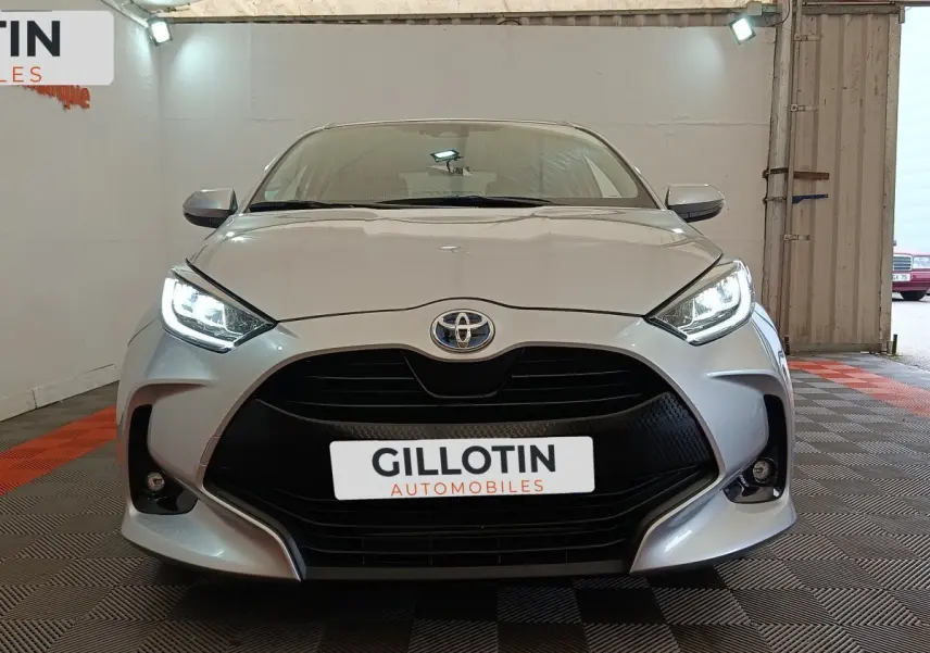 Vue frontale d'une Toyota Yaris Hybride 116h Design gris clair avec phares LED allumés dans un showroom.
