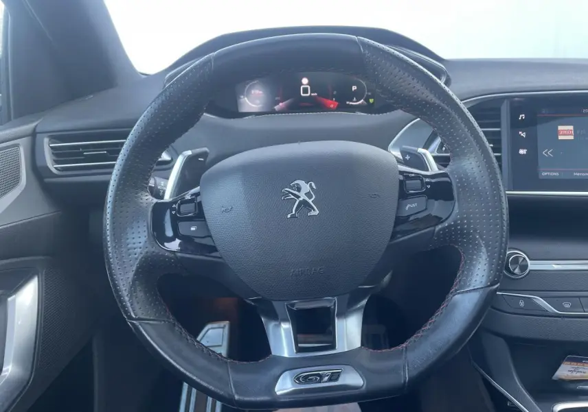 Vue rapprochée du volant cuir perforé noir avec logo Peugeot et badge GT du tableau de bord intérieur anthracite.