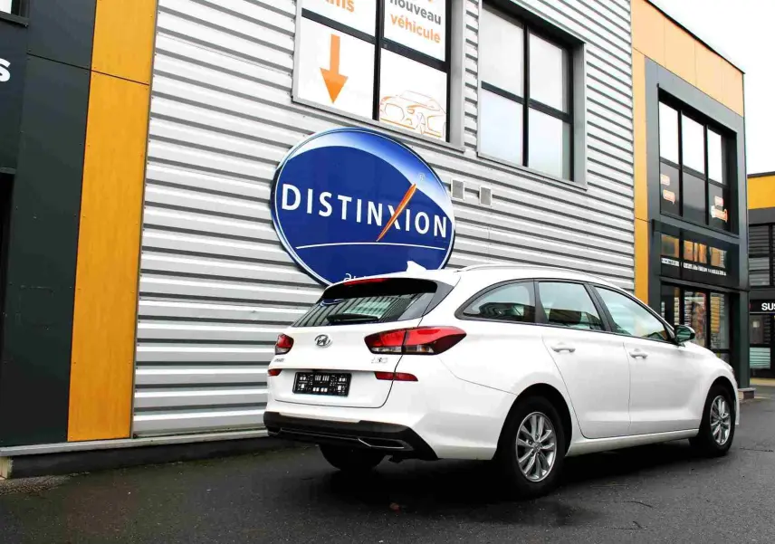 Vue 3/4 arrière droite d'une Hyundai i30 SW blanche polaire garée devant un bâtiment avec un panneau Distinxion.