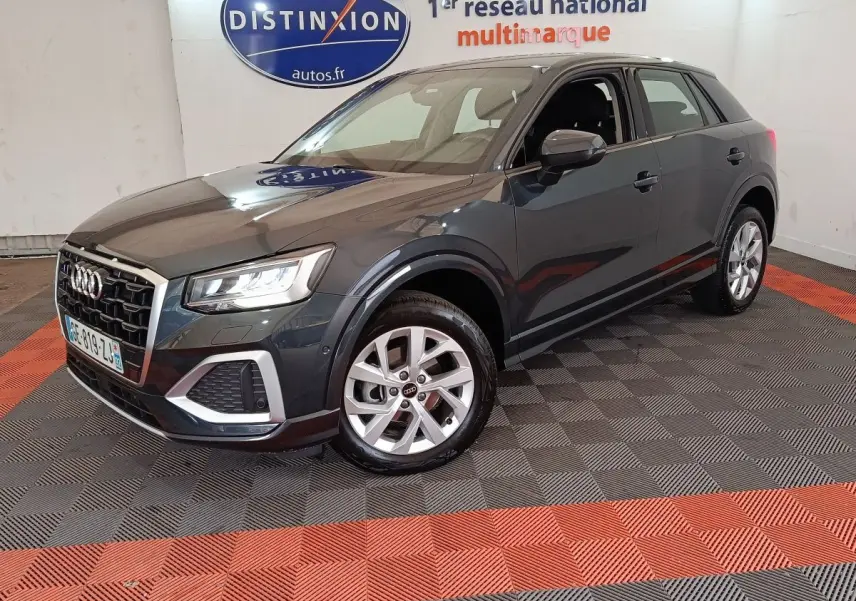 Audi Q2 gris foncé vue 3/4 avant droit, avec jantes alliage et phares LED allumés en intérieur showroom.