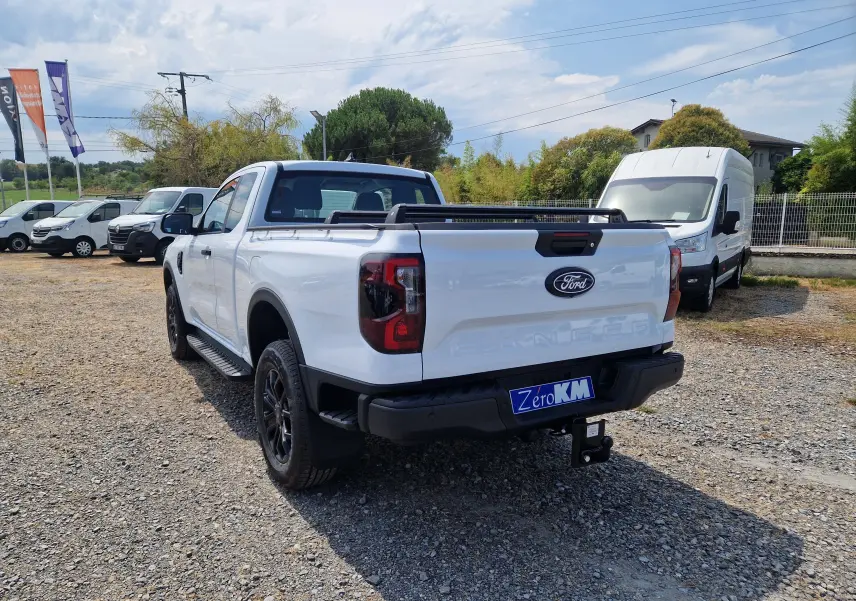 Ford Ranger Super Cabine blanc vu 3/4 arrière droit, avec rideau rigide coulissant et attelage visible sur terrain gravel.
