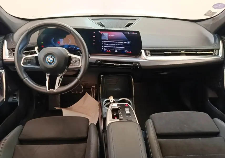 Intérieur BMW X1 xDrive25e M Sport 2023 vu de face, volant cuir alu, écran tactile large, sièges noirs en alcantara.