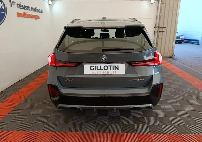 Vue arrière d’un BMW X1 xDrive25e M Sport gris foncé avec feux LED et spoiler de toit visible en intérieur showroom.