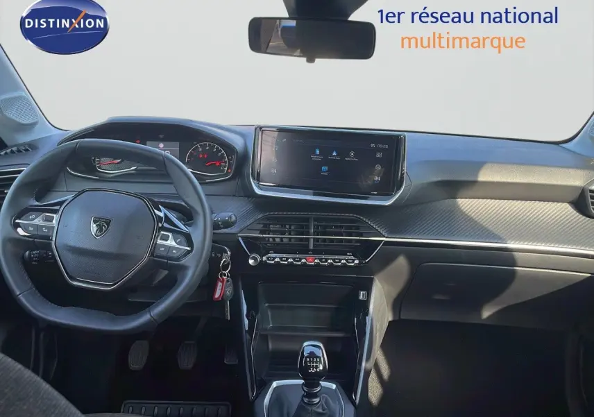 Vue intérieure avant du tableau de bord de la Peugeot 208 blanc banquise 2024 avec volant multifonction et boîte manuelle.