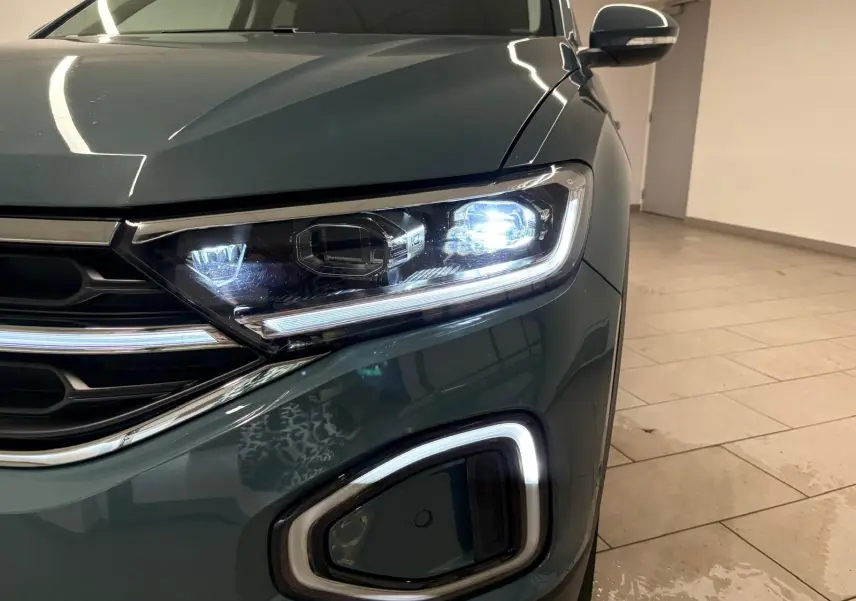 Gros plan sur l'avant droit du Volkswagen T-Roc bleu pétrole 2025, mettant en valeur le phare LED allumé et la calandre.