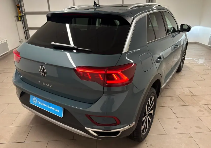Vue 3/4 arrière droite du Volkswagen T-Roc bleu pétrole 2025 avec feux arrière allumés dans un garage.