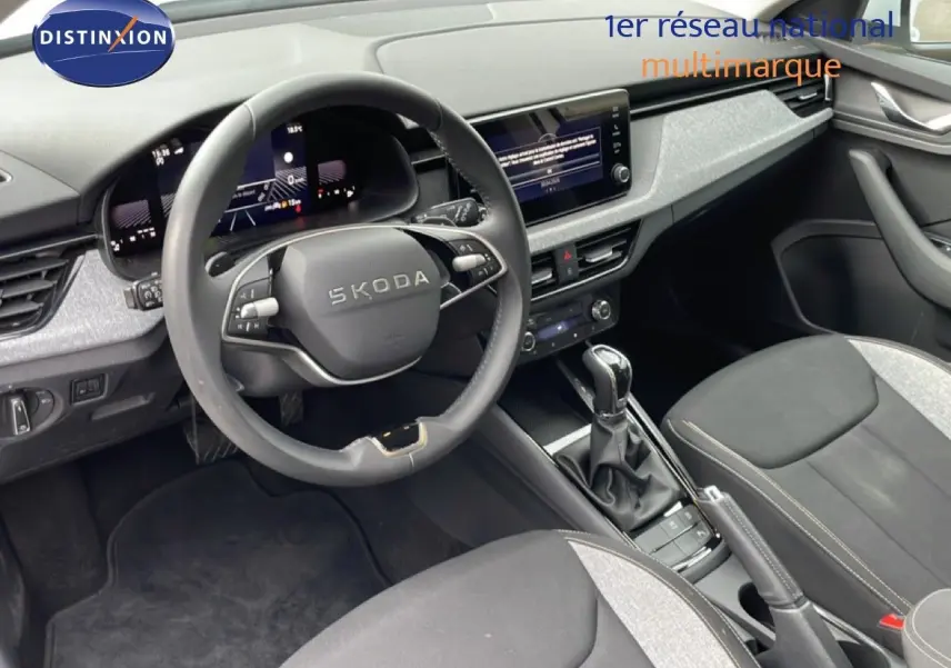 Vue intérieure de face du tableau de bord et volant noir du Skoda Kamiq blanc glacier 2025 avec écran tactile et sellerie tissu gris.