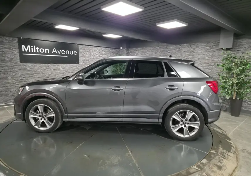 Audi Q2 gris Daytona en vue profil droit dans un showroom avec jantes alliage 18 pouces et vitres surteintées.