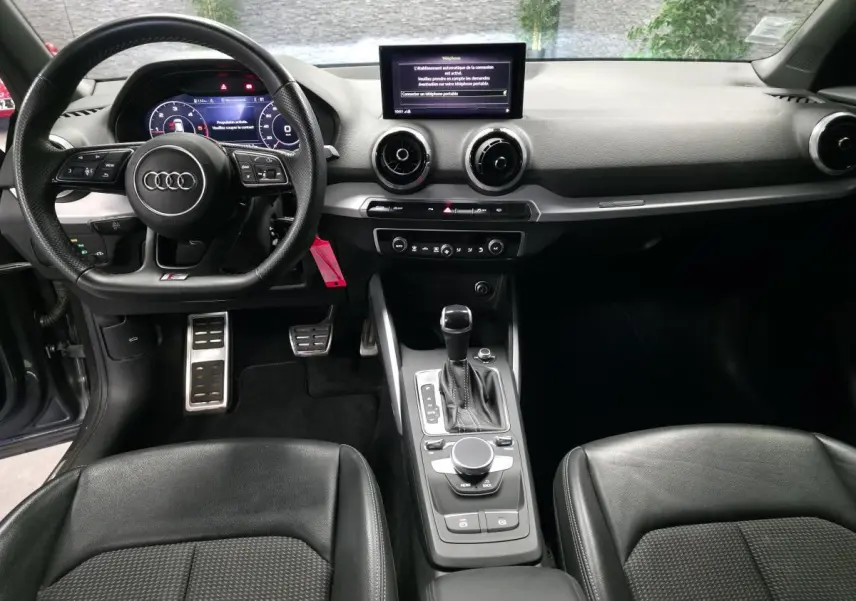 Vue intérieure avant de l'Audi Q2 S line Plus 2023, tableau de bord noir avec volant sport multifonction et console centrale moderne.