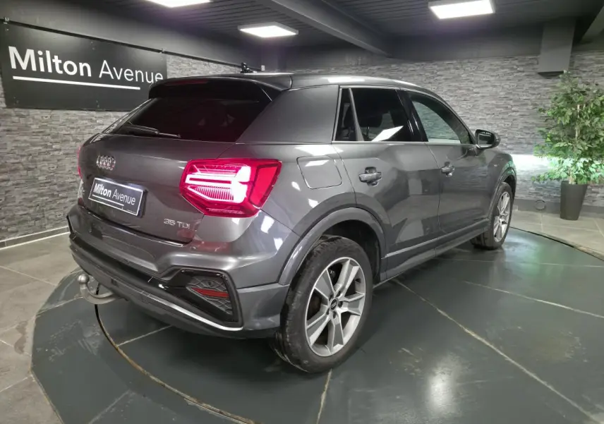 Audi Q2 gris en 3/4 arrière droit, avec feux LED allumés et attelage amovible visible au pare-chocs.