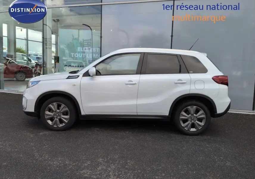 Profil côté gauche d'un Suzuki Vitara blanc avec jantes alu, vitres teintées et lignes de carrosserie marquées.