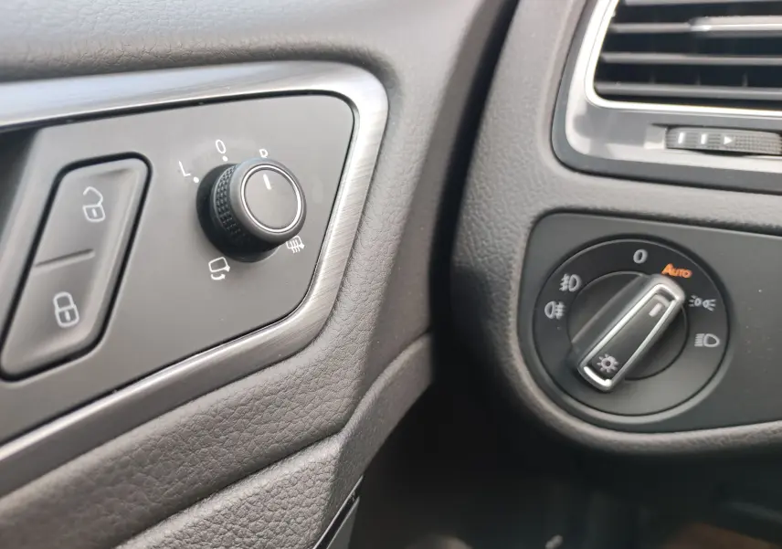 Gros plan sur les commandes d’éclairage et de réglage des rétroviseurs côté gauche dans l’habitacle noir d’une Volkswagen Golf blanche.
