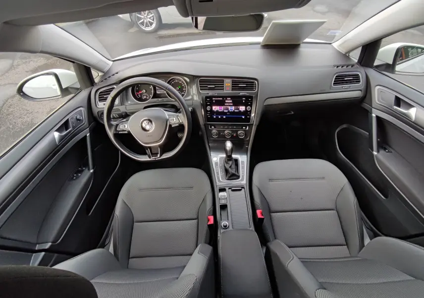 Vue intérieure avant de la Volkswagen Golf 1.5 TSI blanche, montrant le tableau de bord, volant multifonction et sièges noirs.