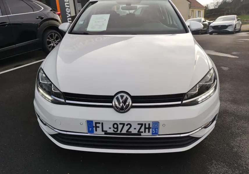 Vue frontale d'une Volkswagen Golf blanche 1.5 TSI EVO 150CV Match de 2019 avec phares LED allumés et plaque d'immatriculation française.