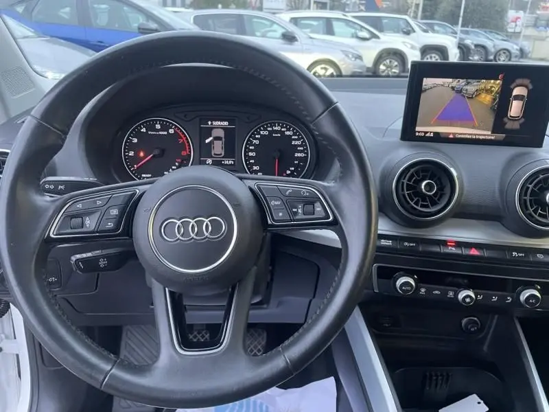 Vue intérieure centrée sur le volant noir d’un Audi Q2 blanc 2020 avec tableau de bord et écran de caméra de recul.