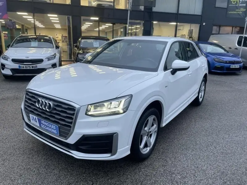 Audi Q2 blanc en 3/4 avant droit, avec calandre noire distinctive et feux LED allumés, stationné devant un showroom.