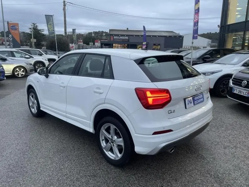 Audi Q2 blanc vue 3/4 arrière droit avec feux allumés, garé en extérieur sur un parking de voitures d'occasion.