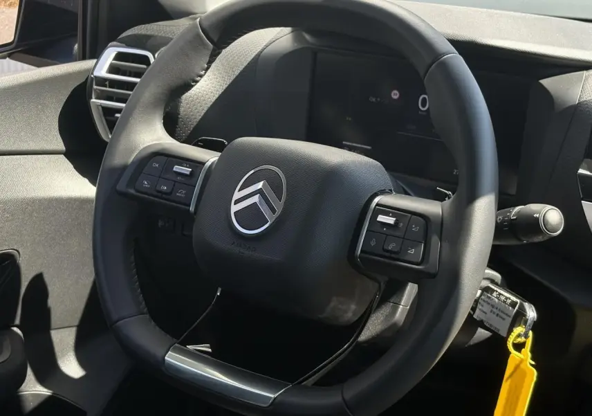 Gros plan sur le volant cuir noir de la Citroën C4 Hybrid 2025, avec commandes intégrées et tableau de bord numérique en arrière-plan.