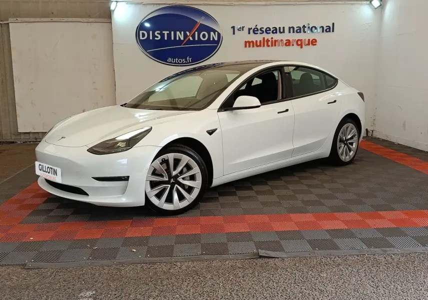 Tesla Model 3 Grande Autonomie blanc vue 3/4 avant droit, stationnée sur sol à damiers rouge et noir en intérieur.