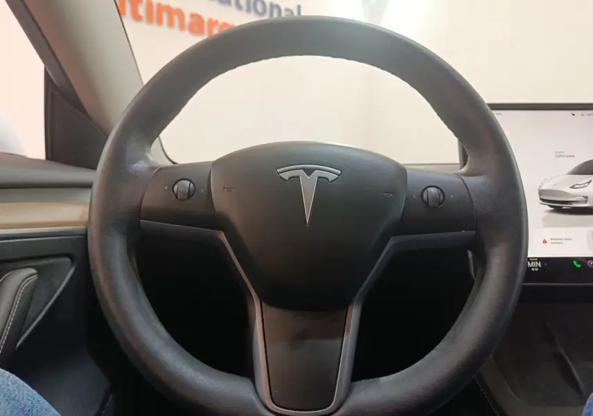 Vue rapprochée du volant en cuir synthétique noir d'une Tesla Model 3 blanche, avec écran tactile visible à droite.