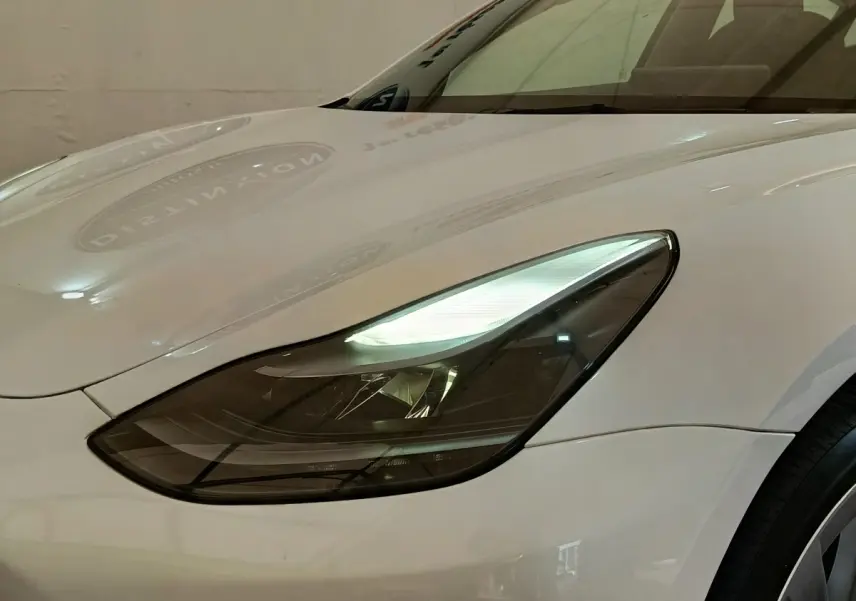 Gros plan sur le phare avant gauche d’une Tesla Model 3 blanche, mettant en valeur son design épuré et moderne.