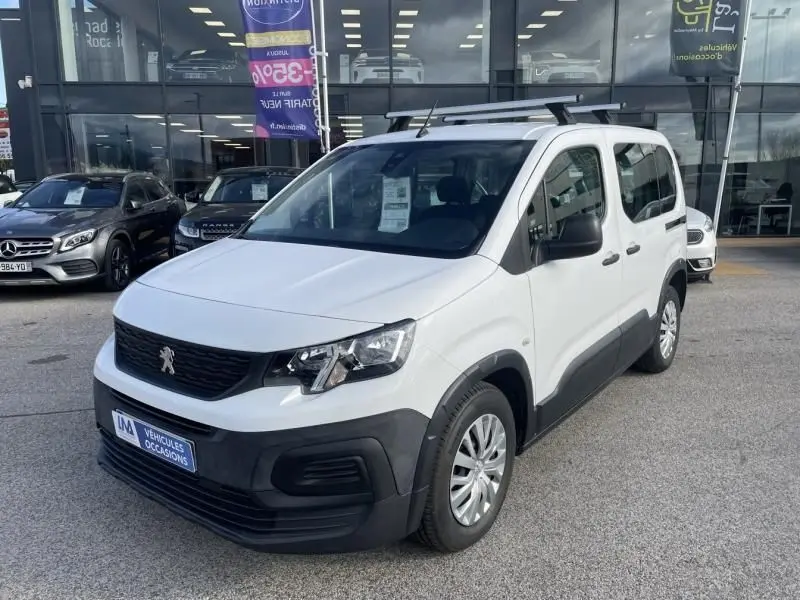 Peugeot Rifter blanc vue 3/4 avant droit, avec protections noires sur les bas de caisse et barres de toit, stationné devant un showroom.
