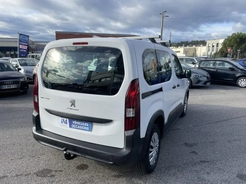 Vue 3/4 arrière droite d'un Peugeot Rifter blanc 2020 avec hayon, barres de toit et attelage visible sur parking.