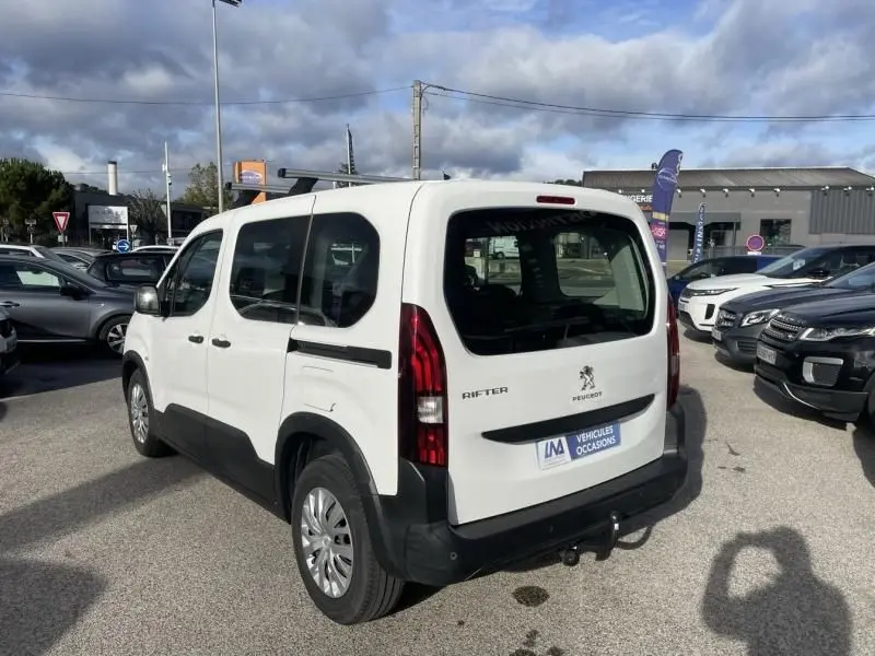 Vue 3/4 arrière droite du Peugeot Rifter blanc 2020 avec hayon et barres de toit visibles sur un parking.