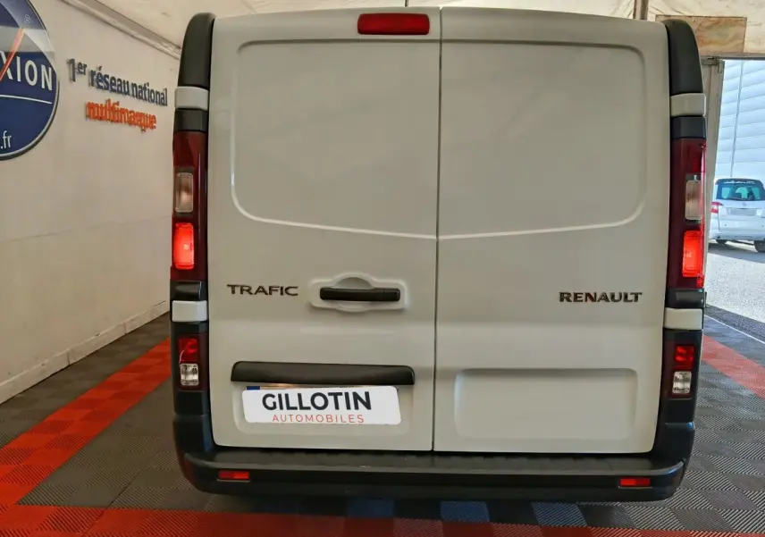 Vue arrière d’un Renault Trafic fourgon blanc avec portes battantes et feux arrière allumés en intérieur showroom.