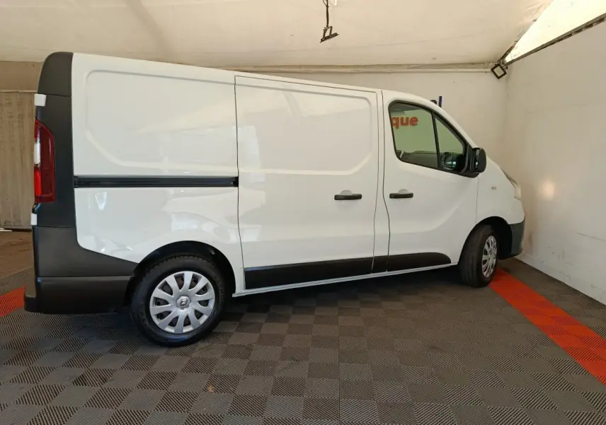 Profil droit d’un utilitaire Renault Trafic blanc avec cloison pleine, stationné sur sol à damier gris et rouge.