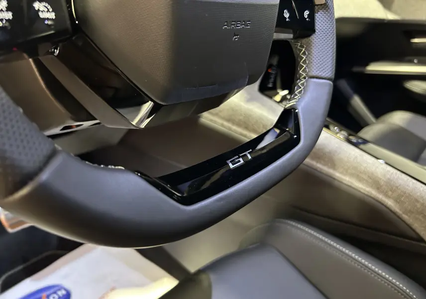 Gros plan sur le volant GT noir perforé de l’intérieur du Peugeot 3008 Hybrid blanc Okenite, avec détails de la console.
