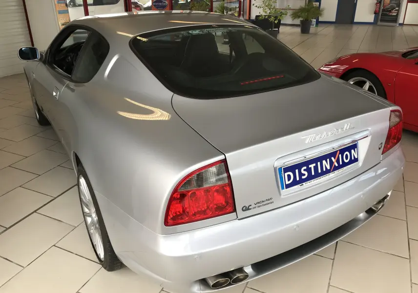 Vue 3/4 arrière droite d'une Maserati Coupé gris clair de 2003 avec double sortie d'échappement et feux arrière rouges.