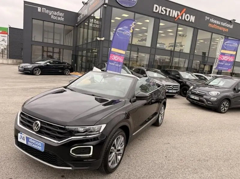 Volkswagen T-Roc Cabriolet noir métal en 3/4 avant, capote ouverte, devant un showroom multimarque.