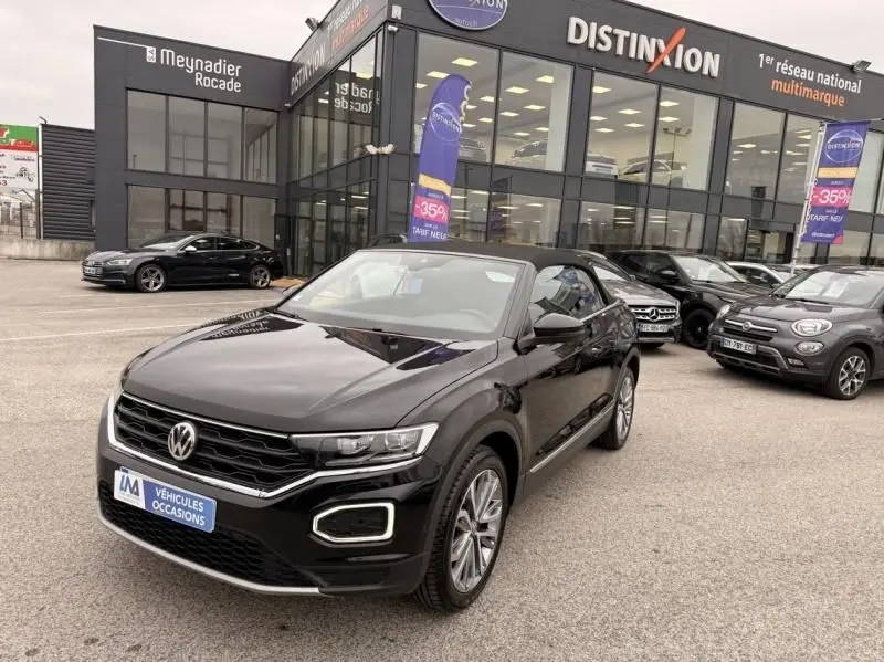 Volkswagen T-Roc Cabriolet noir métal vu en 3/4 avant, capote ouverte, devant un showroom multimarque.