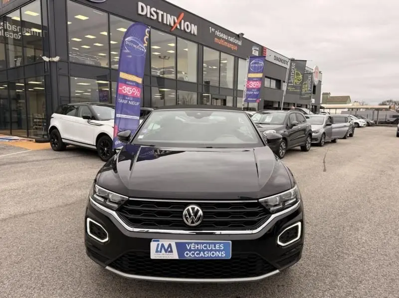 Volkswagen T-Roc Cabriolet noir métal vue de face, capote ouverte, devant un concessionnaire multimarque.