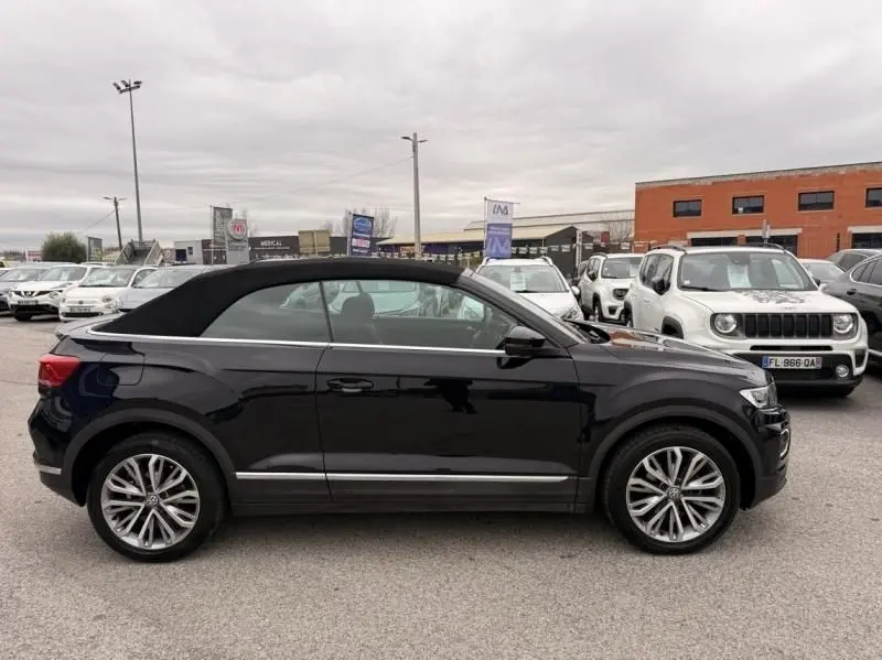 Vue de profil côté gauche d’un Volkswagen T-Roc Cabriolet noir métal avec toit fermé, stationné en extérieur.