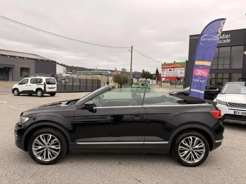 Vue de profil côté gauche d'un Volkswagen T-Roc Cabriolet noir métal 2020 avec toit ouvert sur parking.