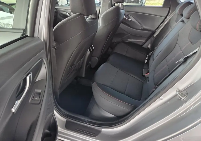 Vue intérieure côté arrière droit de la Hyundai i30 SW N-LINE 2025, sièges tissu noirs avec surpiqûres rouges, porte gris foncé ouverte.